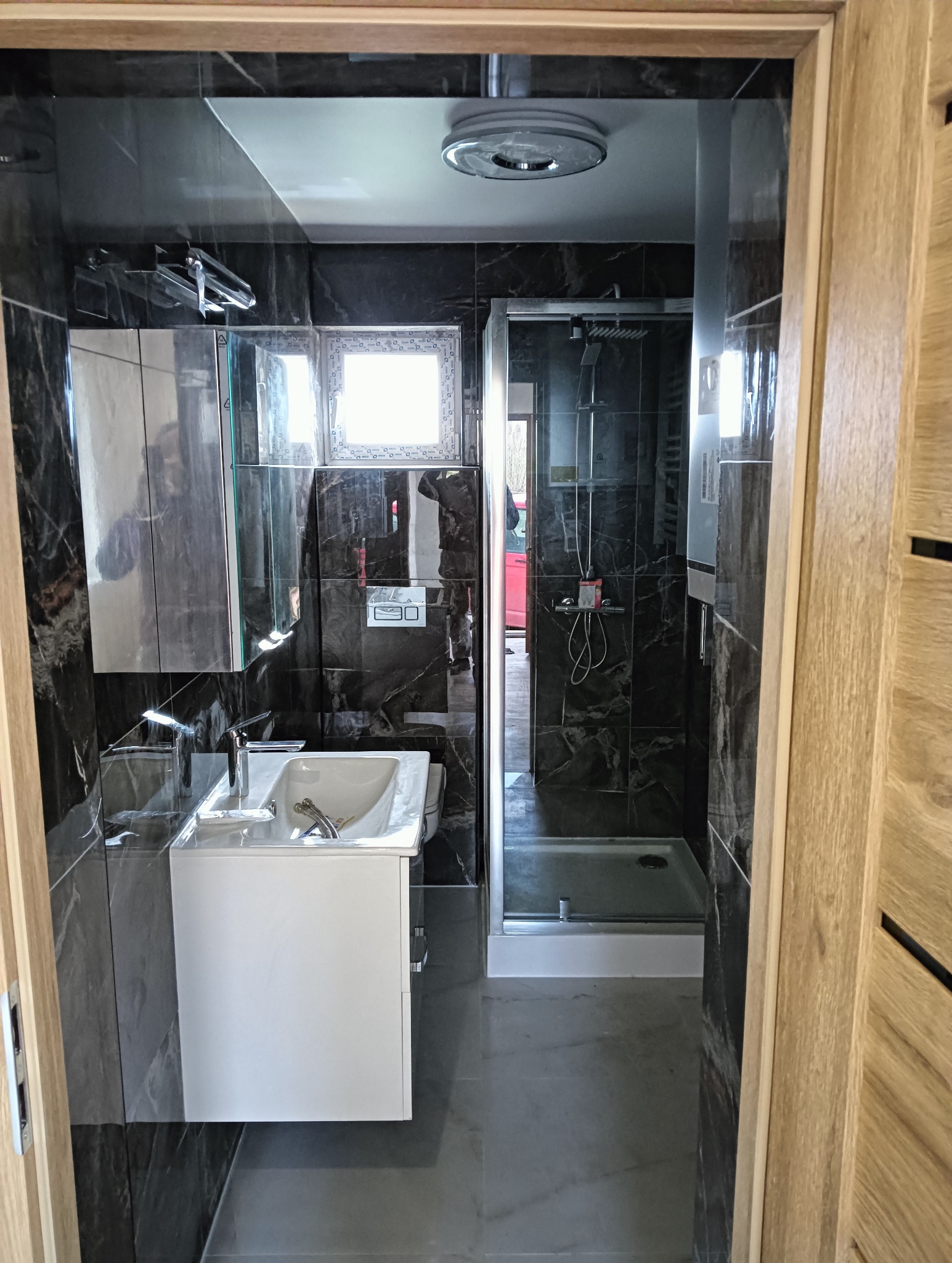 Wykończona łazienka z kabiną prysznicową i umywalką na szafce, wyłożona czarnymi płytkami imitującymi marmur. Widok zza drewnianej framugi drzwi.
