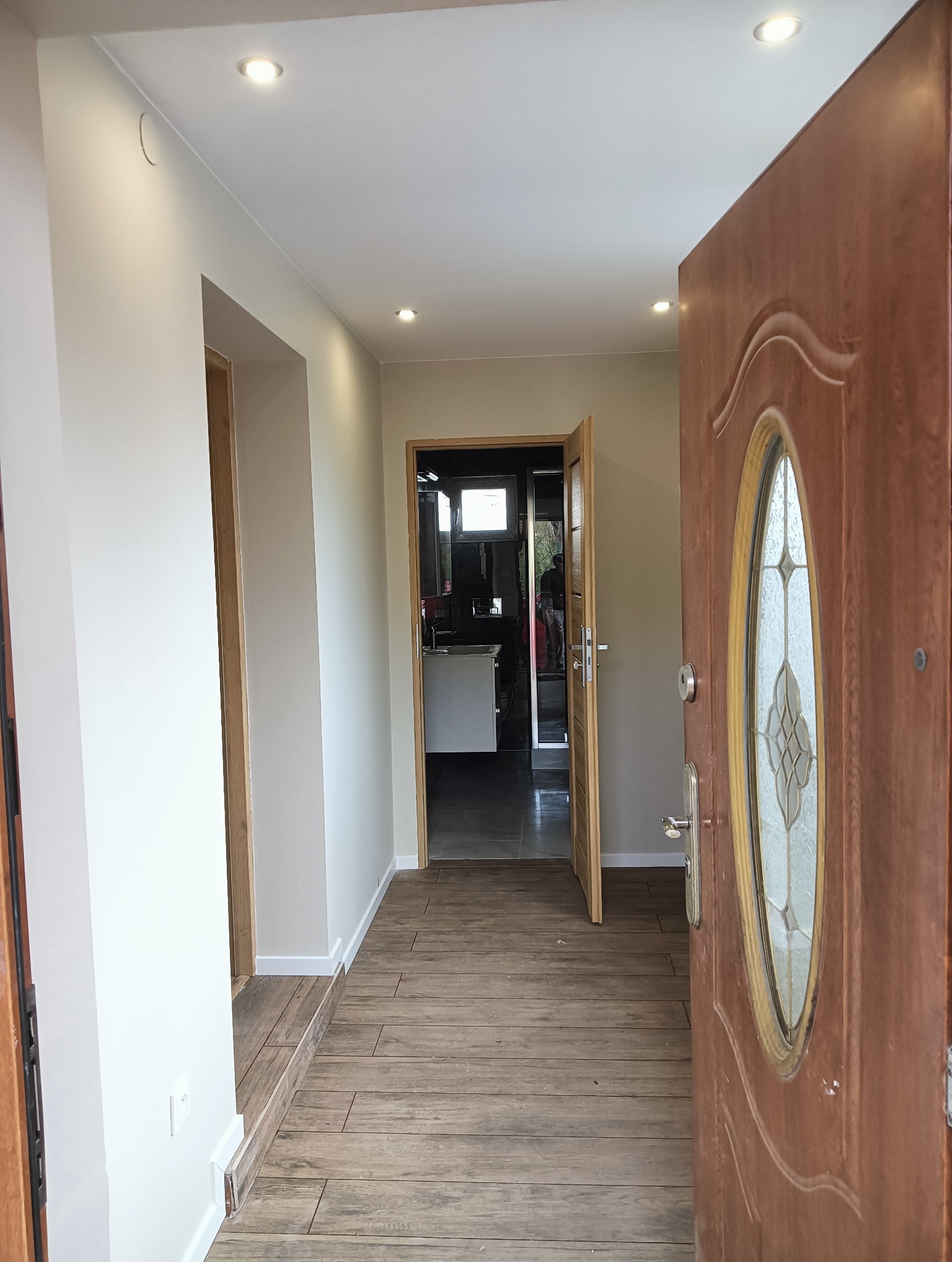 Korytarz po remoncie z drewnopodobną podłogą. Widok przez otwarte drzwi wejściowe na wnętrze z oświetleniem punktowym i jasnymi ścianami. W tle widoczna łazienka.