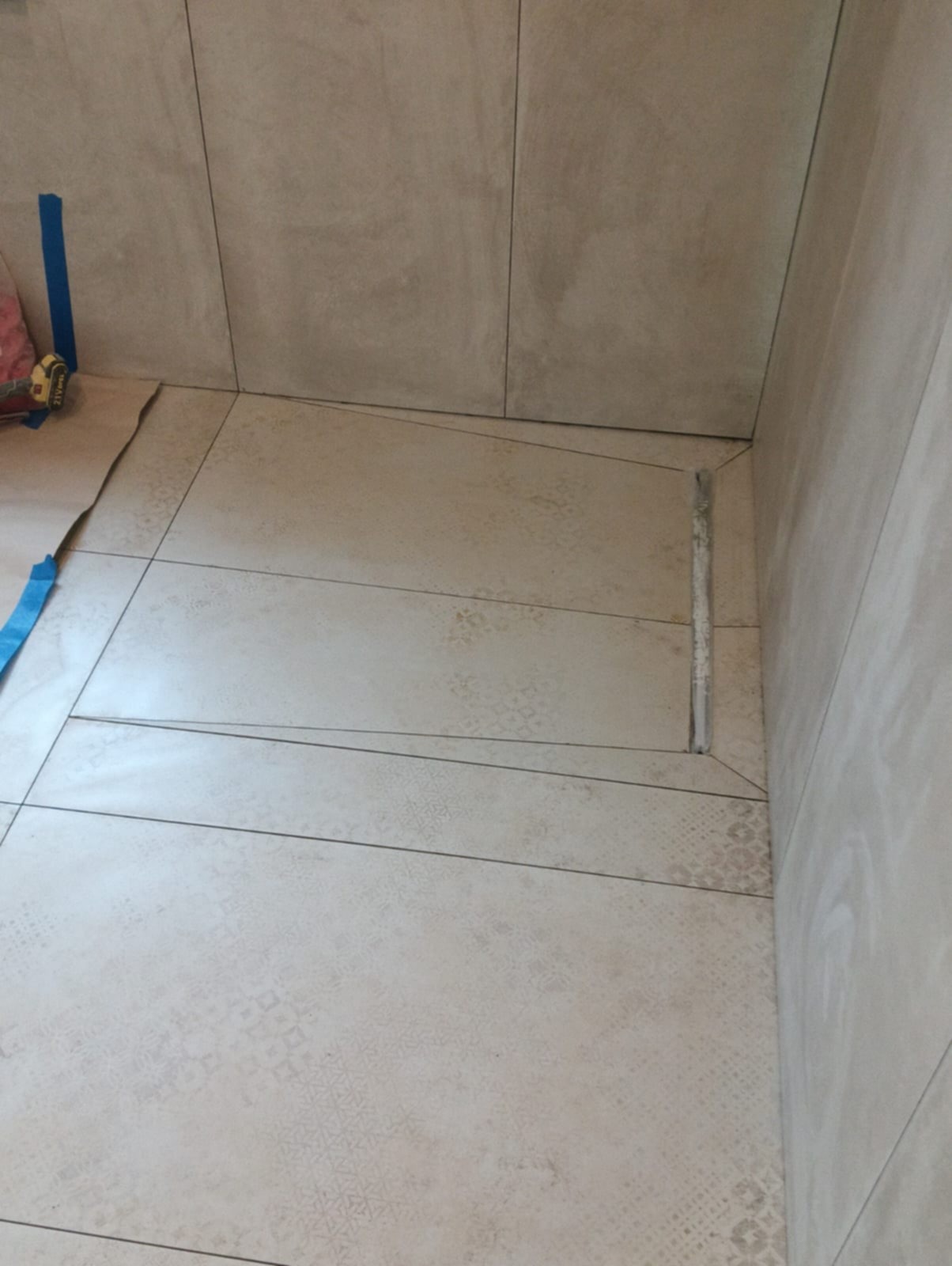 Ułożone płytki podłogowe i ścienne w łazience, z widocznym odpływem liniowym. Fragment podłogi zabezpieczony kartonem i taśmą malarską. Jasne, beżowe odcienie płytek.