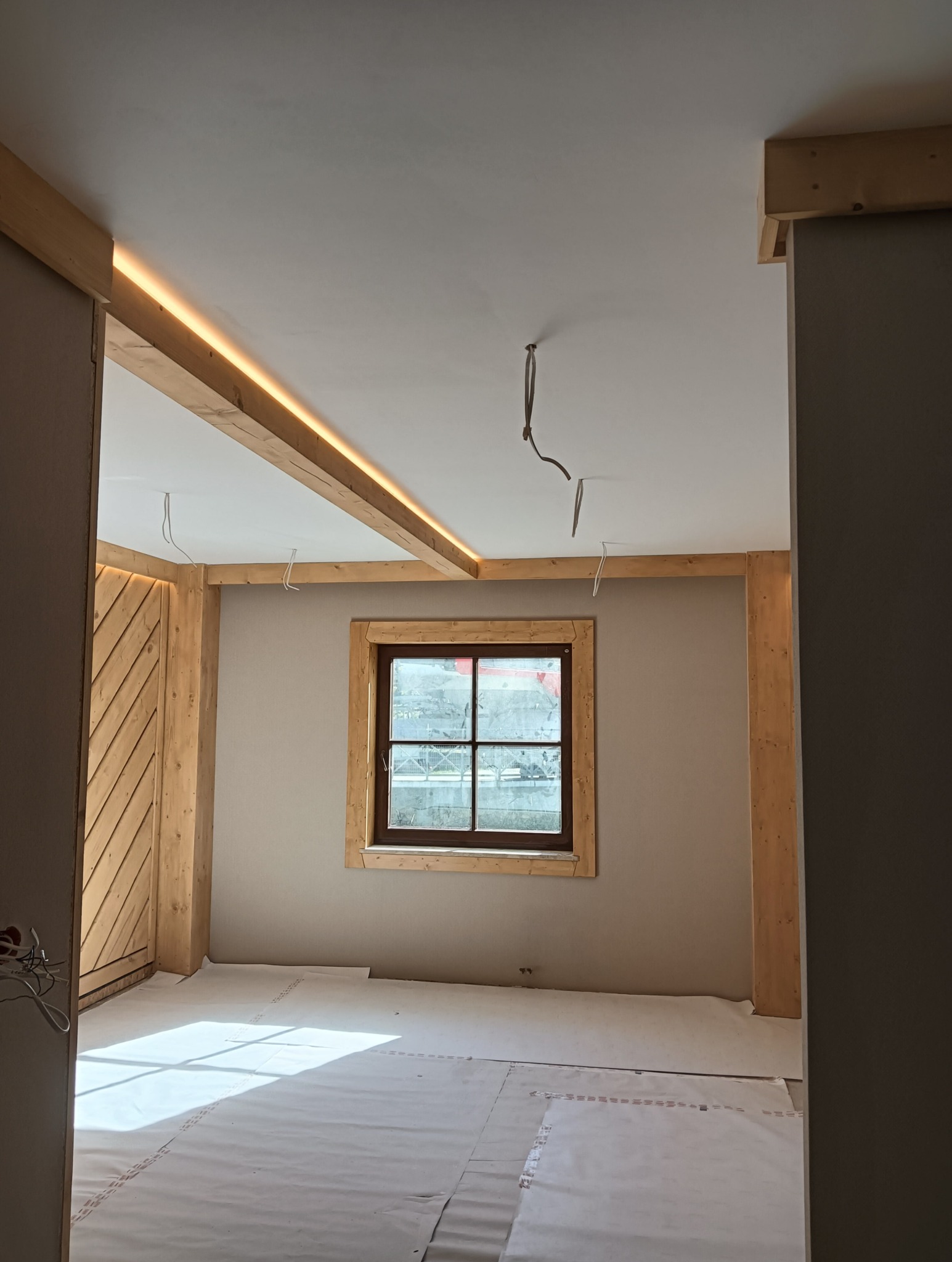 Pokój w trakcie wykończenia: drewniane belki z oświetleniem LED, okno w drewnianej ramie, zabezpieczona podłoga. Widoczne kable wystające z sufitu.