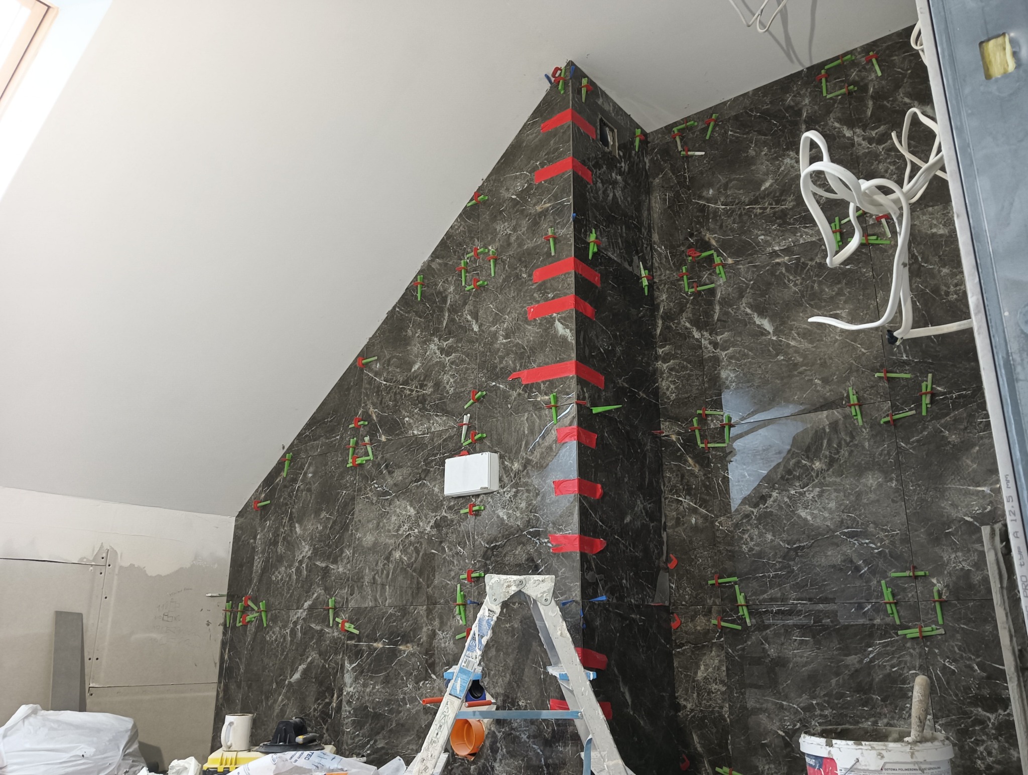 Ściana w łazience wyłożona ciemnymi płytkami imitującymi marmur, z użyciem poziomowania krzyżykami i taśmy. Widoczna drabina i narzędzia glazurnicze.