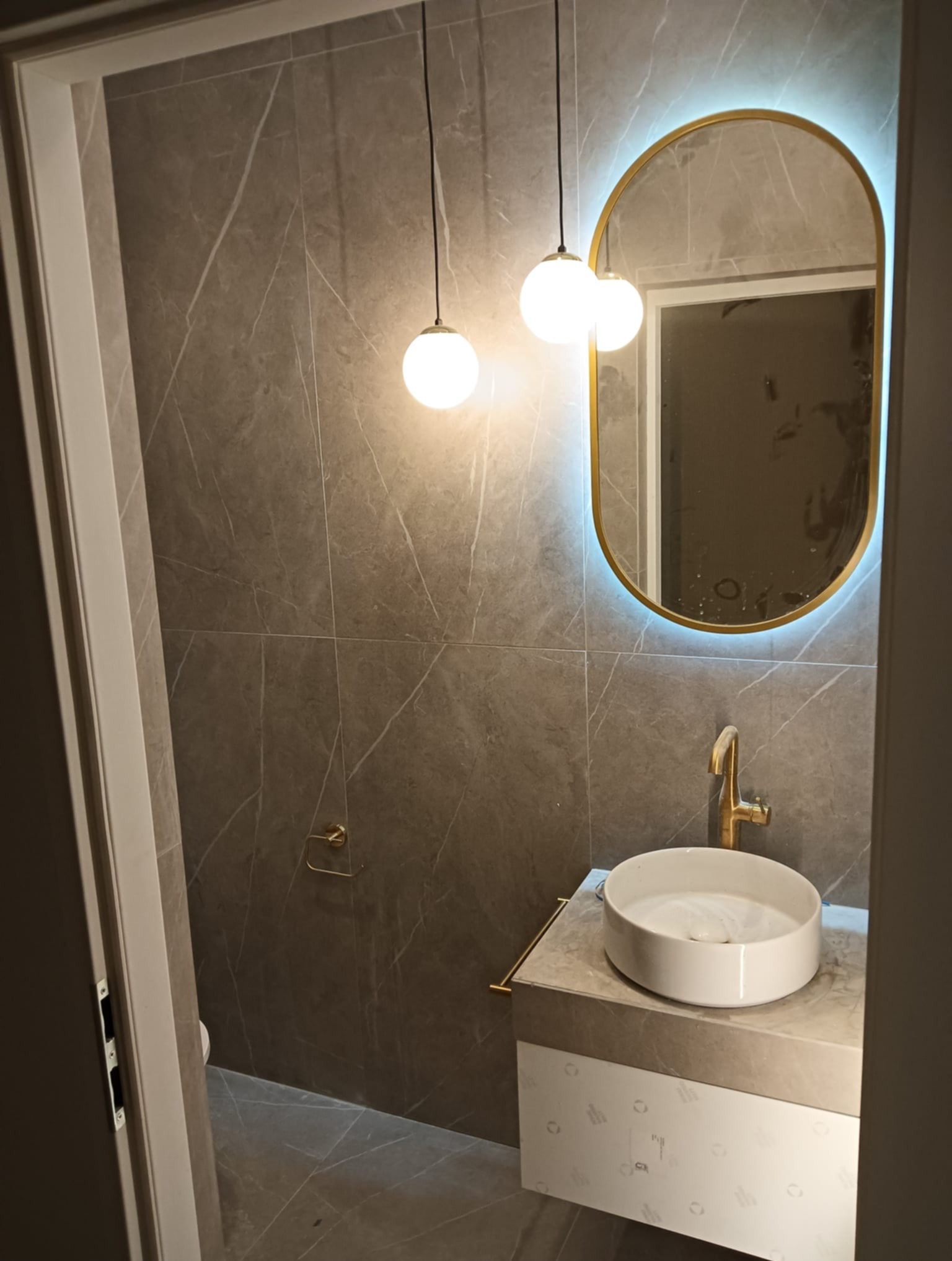 Nowoczesna łazienka z szarymi płytkami imitującymi marmur, okrągłą umywalką nablatową i podświetlanym lustrem w złotej ramie. Minimalistyczne oświetlenie.