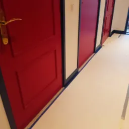 Korytarz z trzema odnowionymi drzwiami w odcieniach czerwieni, z widoczną taśmą malarską na podłodze zabezpieczającą przed zabrudzeniem. Złota klamka na pierwszej parze drzwi.