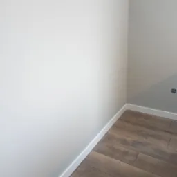 Wykończony narożnik pokoju z ułożonymi panelami podłogowymi w odcieniach brązu i pomalowanymi na biało ścianami z listwami przypodłogowymi.