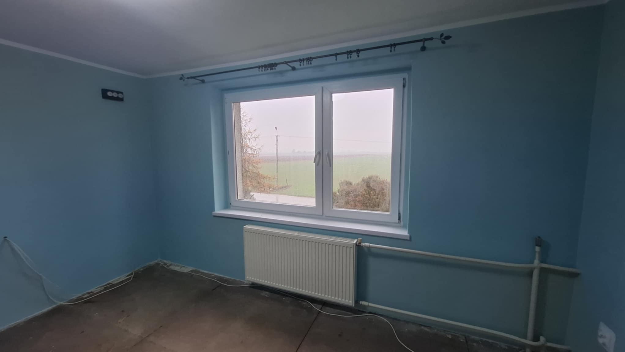 Pomalowane na niebiesko wnętrze pokoju z oknem, grzejnikiem i odsłoniętą podłogą przed położeniem paneli.