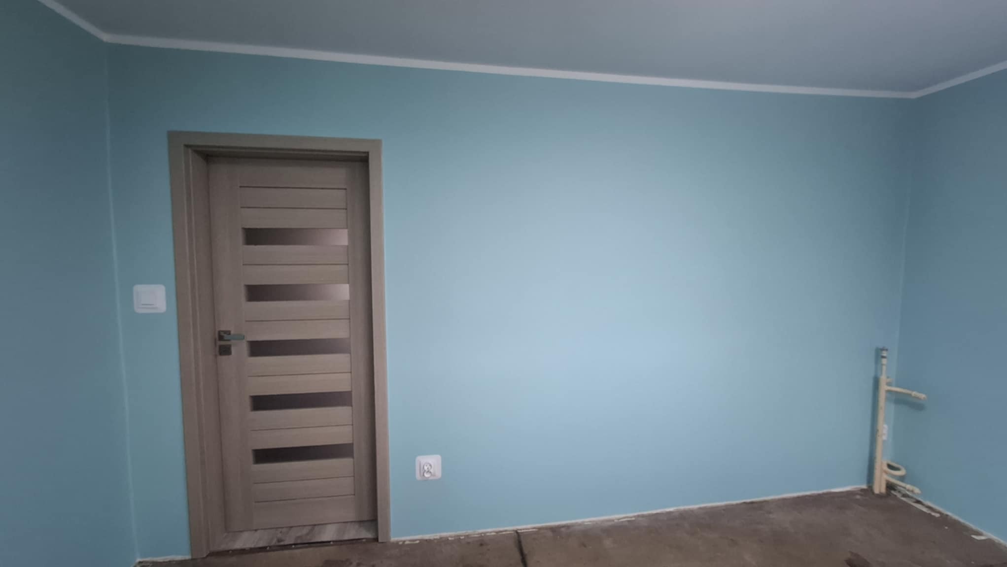 Świeżo pomalowane na błękitno wnętrze pokoju z zamontowaną, nowoczesną, drewnianą ościeżnicą z poziomymi przeszkleniami. Na podłodze widoczne ślady po remoncie. W rogu wystają rury instalacji wodnej.