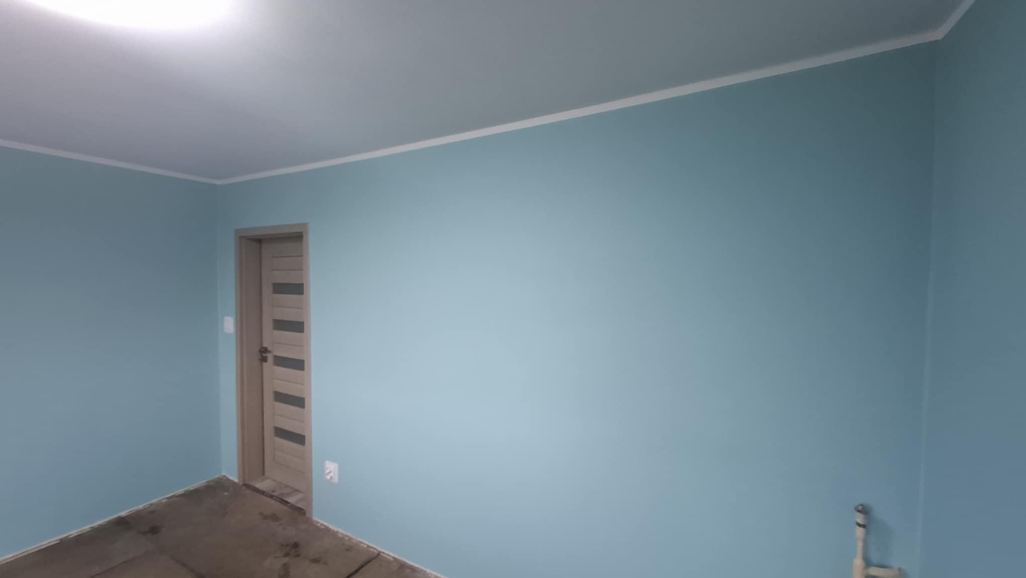 Świeżo pomalowane na błękitno ściany pokoju z białą listwą pod sufitem, widoczna drewniana ościeżnica drzwiowa z przeszklonymi panelami oraz surowa, betonowa podłoga przed ułożeniem paneli.