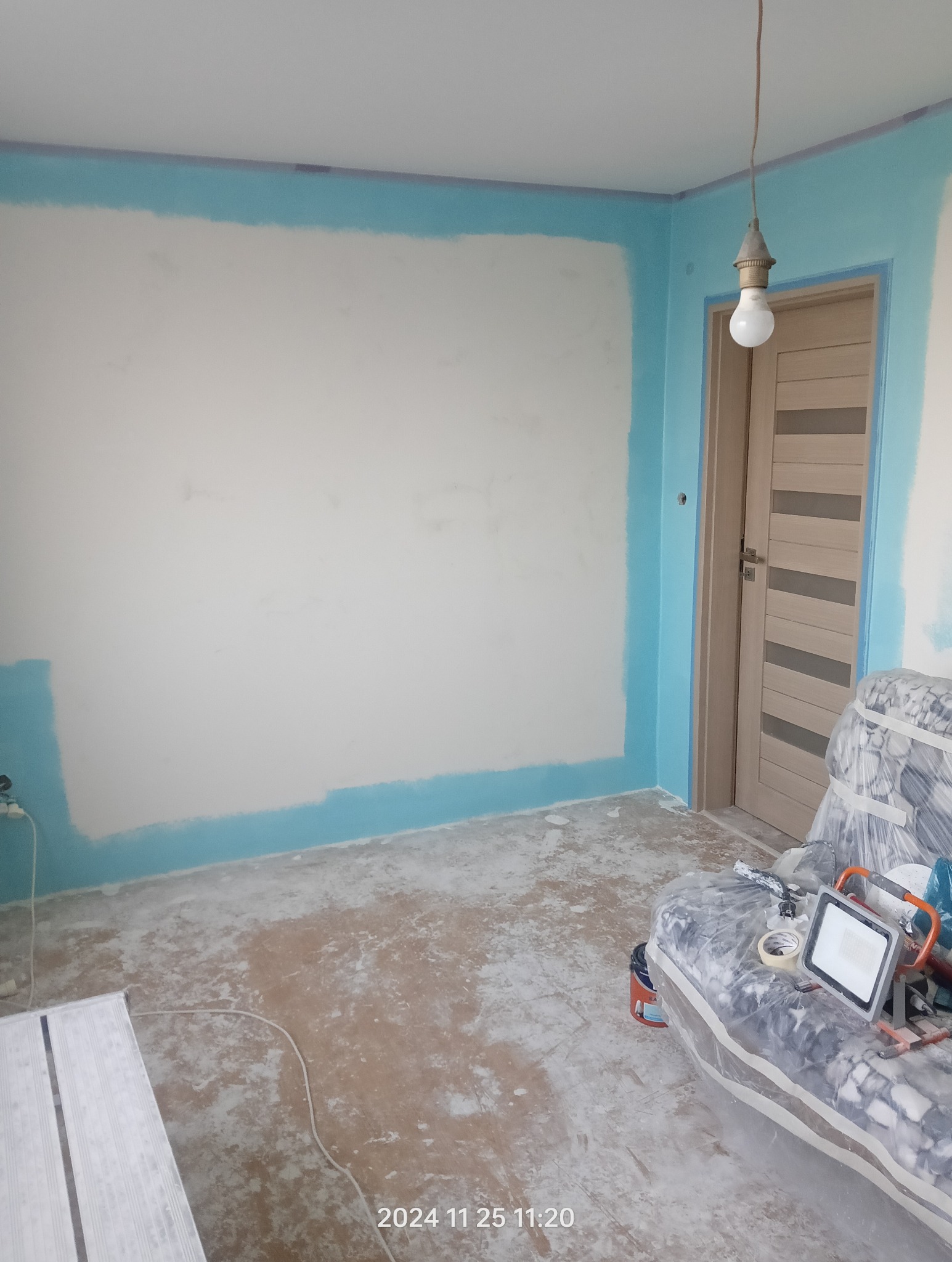 Pokój w trakcie przygotowania do malowania, z niebieską farbą na obrzeżach ścian i sufitu, zabezpieczony mebel folią, widoczne narzędzia i lampa halogenowa.