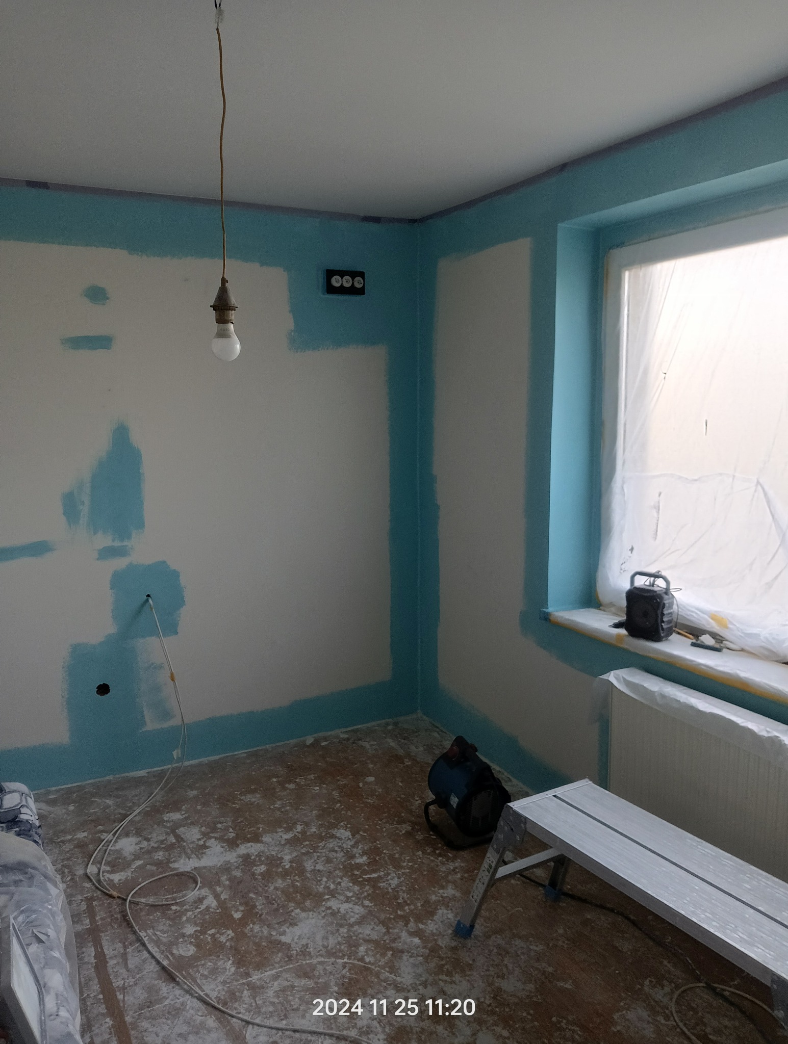 Pokój w trakcie prac przygotowawczych do malowania: ściany częściowo zabezpieczone taśmą malarską i pokryte podkładem, widoczne ślady po taśmie, podłoga zakurzona, drabina aluminiowa, wisząca...