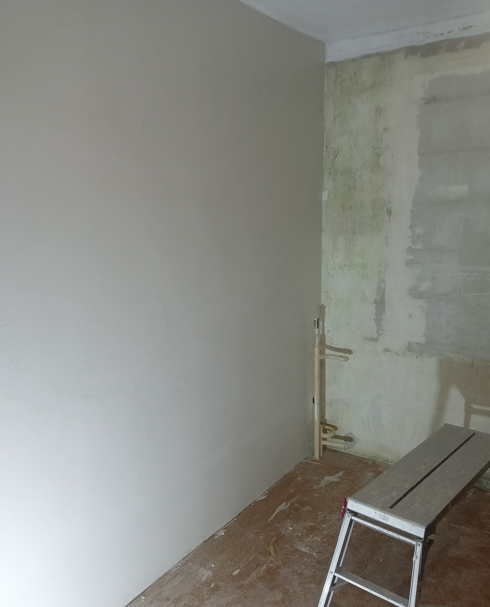 Ściana po nałożeniu gładzi, widoczna drabina malarska, fragment instalacji hydraulicznej przy narożniku ścian. Jedna ściana wygładzona, druga w trakcie przygotowania do prac wykończeniowych.