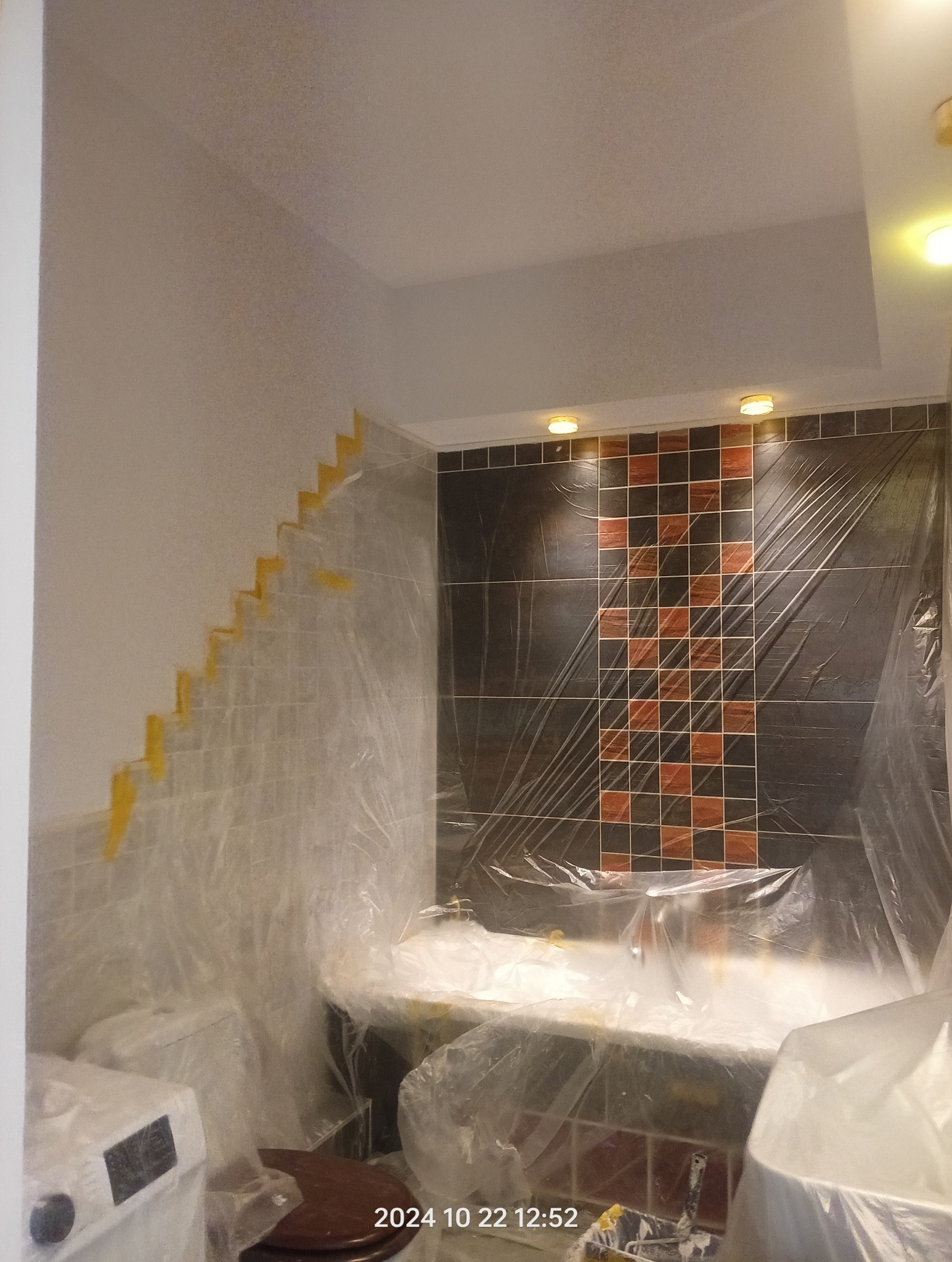 Łazienka w trakcie zabezpieczania folią malarską przed pracami wykończeniowymi, widoczne taśmy malarskie na ścianie, płytki częściowo zasłonięte folią.