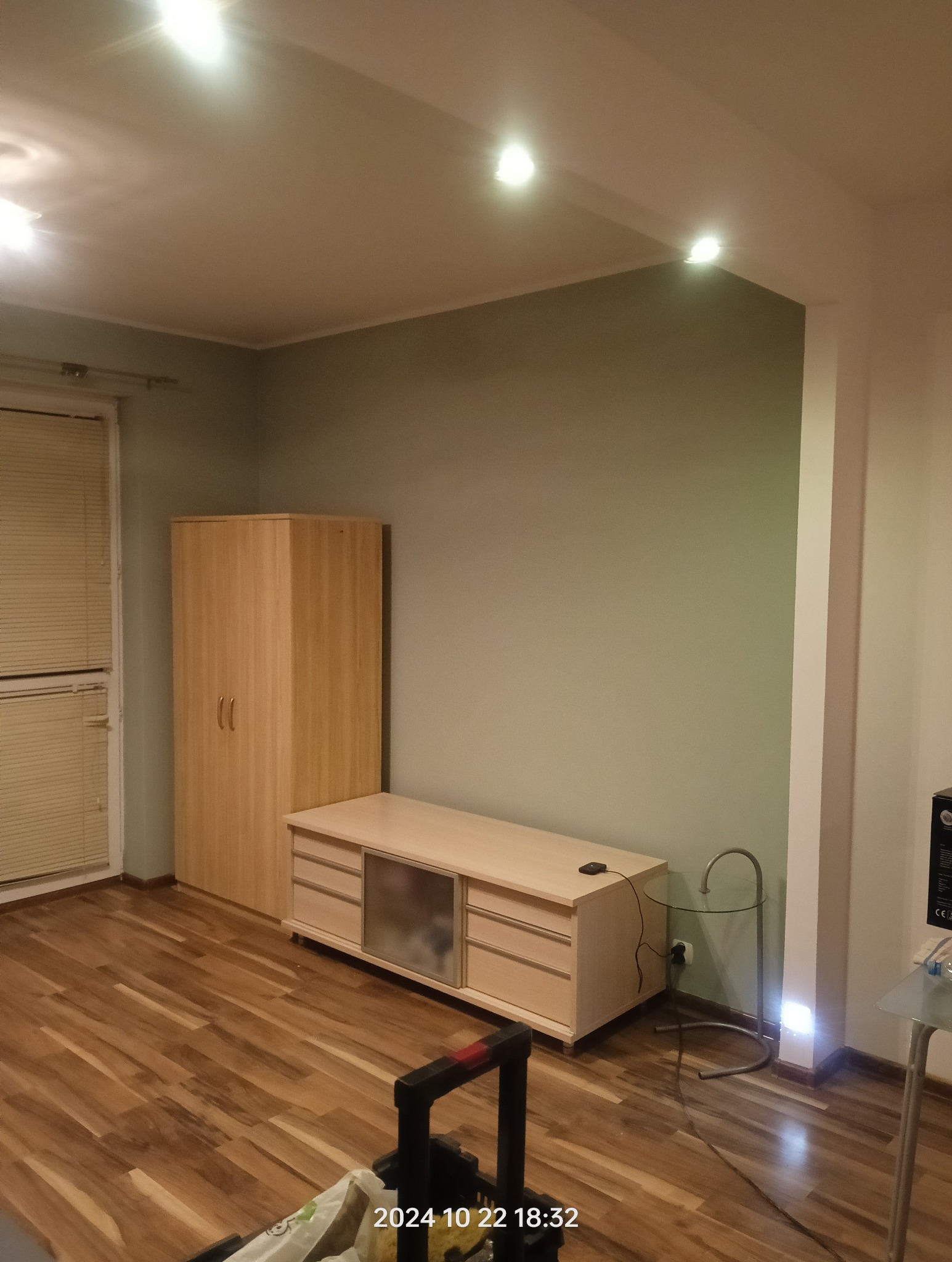 Pokój po malowaniu ścian na kolor oliwkowy, z widocznymi meblami w odcieniach drewna i świeżo ułożonymi panelami podłogowymi. W dolnej części kadru widoczny wózek z narzędziami.