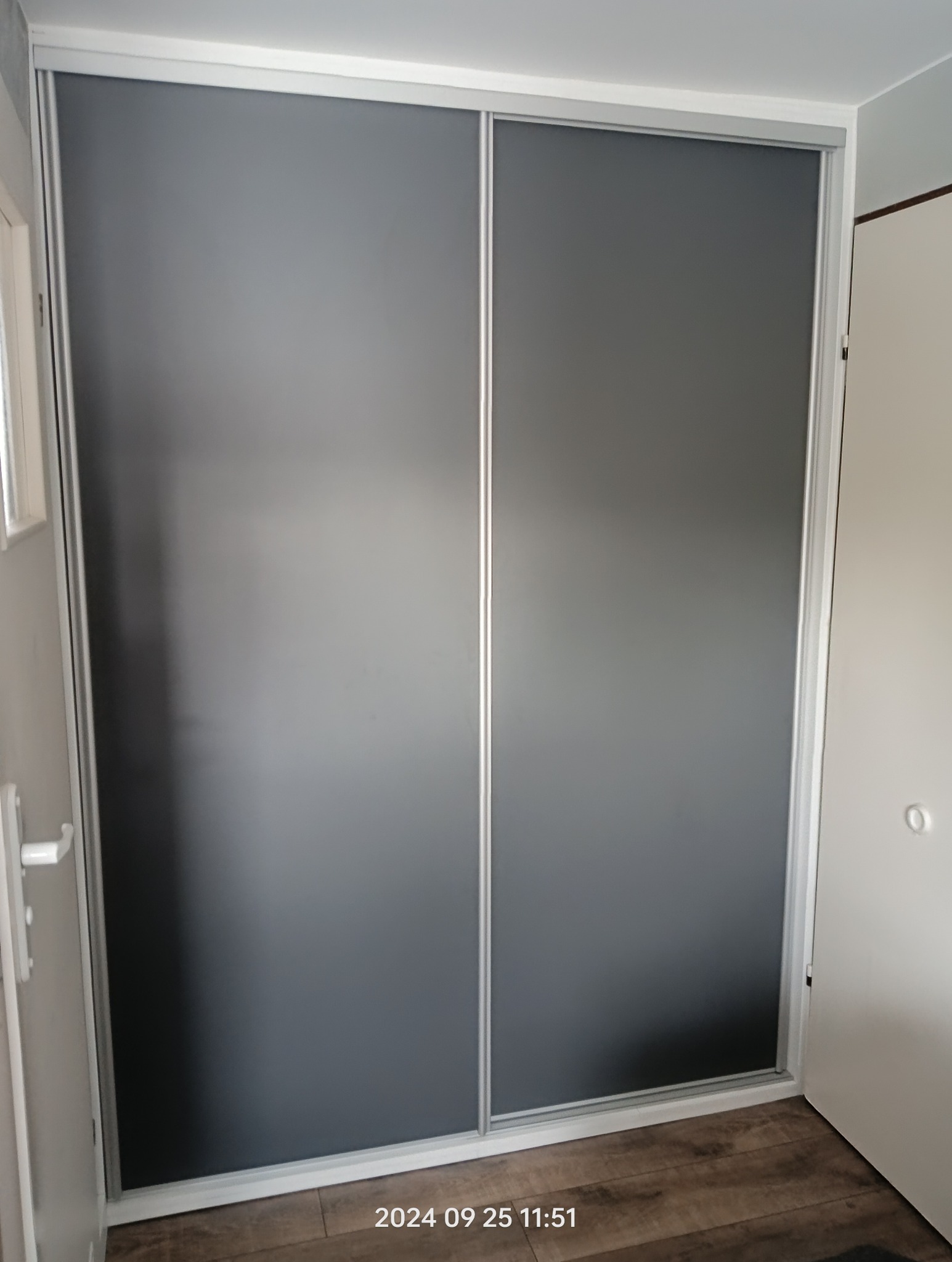 Nowoczesna szafa wnękowa z przesuwnymi, szarymi drzwiami i srebrnymi ramami, widoczna w rogu pokoju z drewnianą podłogą.