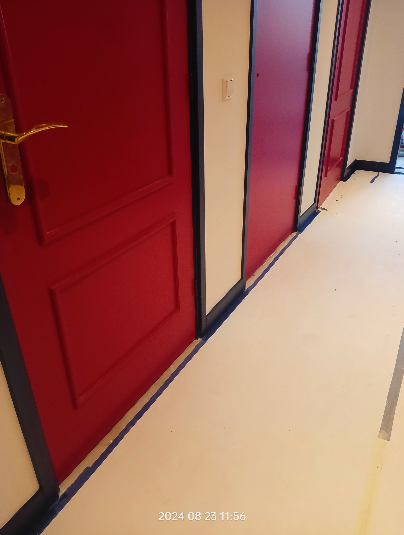 Korytarz z trzema odnowionymi drzwiami w odcieniach czerwieni, z widoczną taśmą malarską na podłodze zabezpieczającą przed zabrudzeniem. Złota klamka na pierwszej parze drzwi.