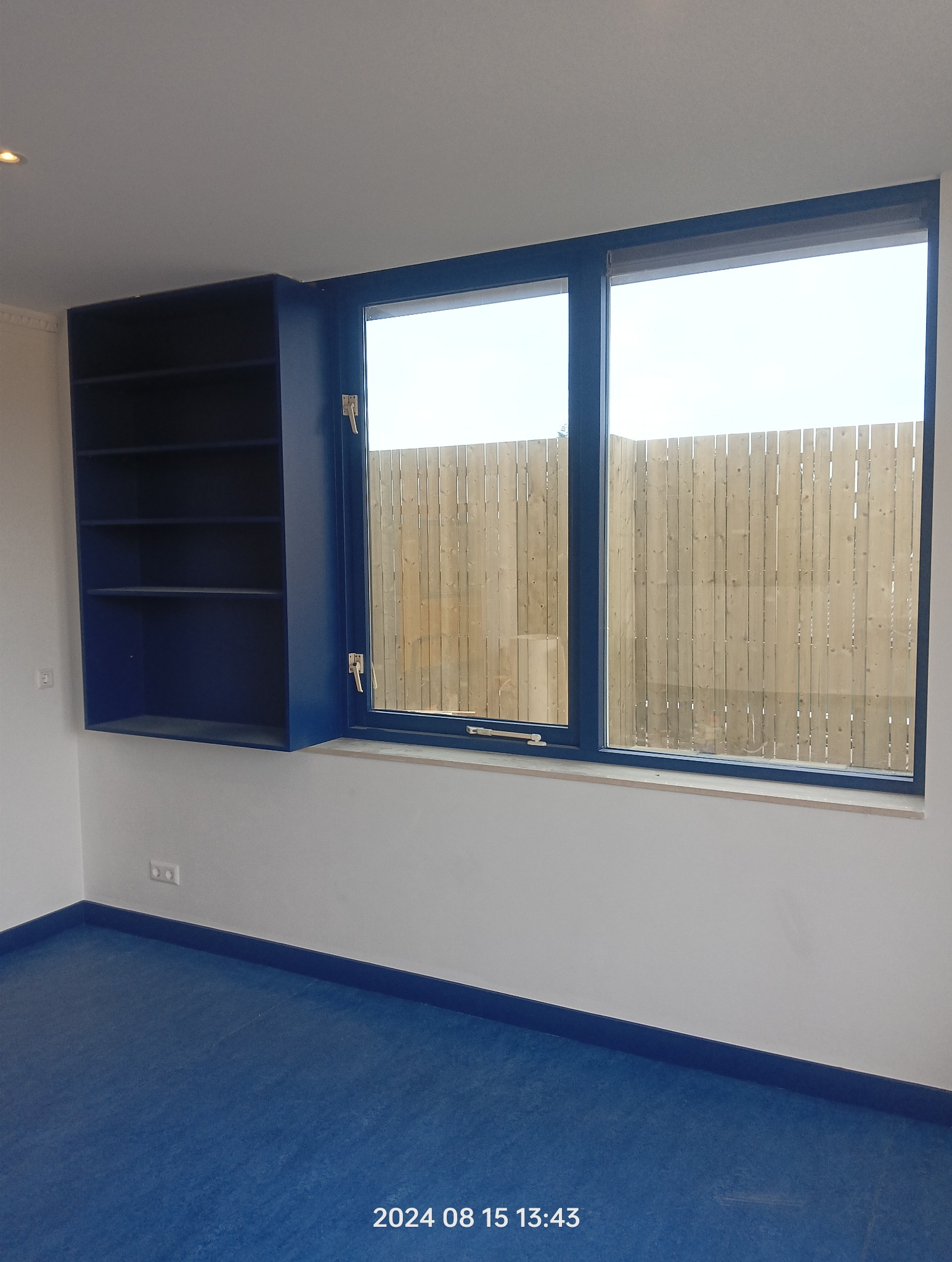 Niebieska zabudowa okienna z szafką wiszącą w nowo wykończonym pokoju z niebieską podłogą i białymi ścianami, widok na drewniany płot za oknem.