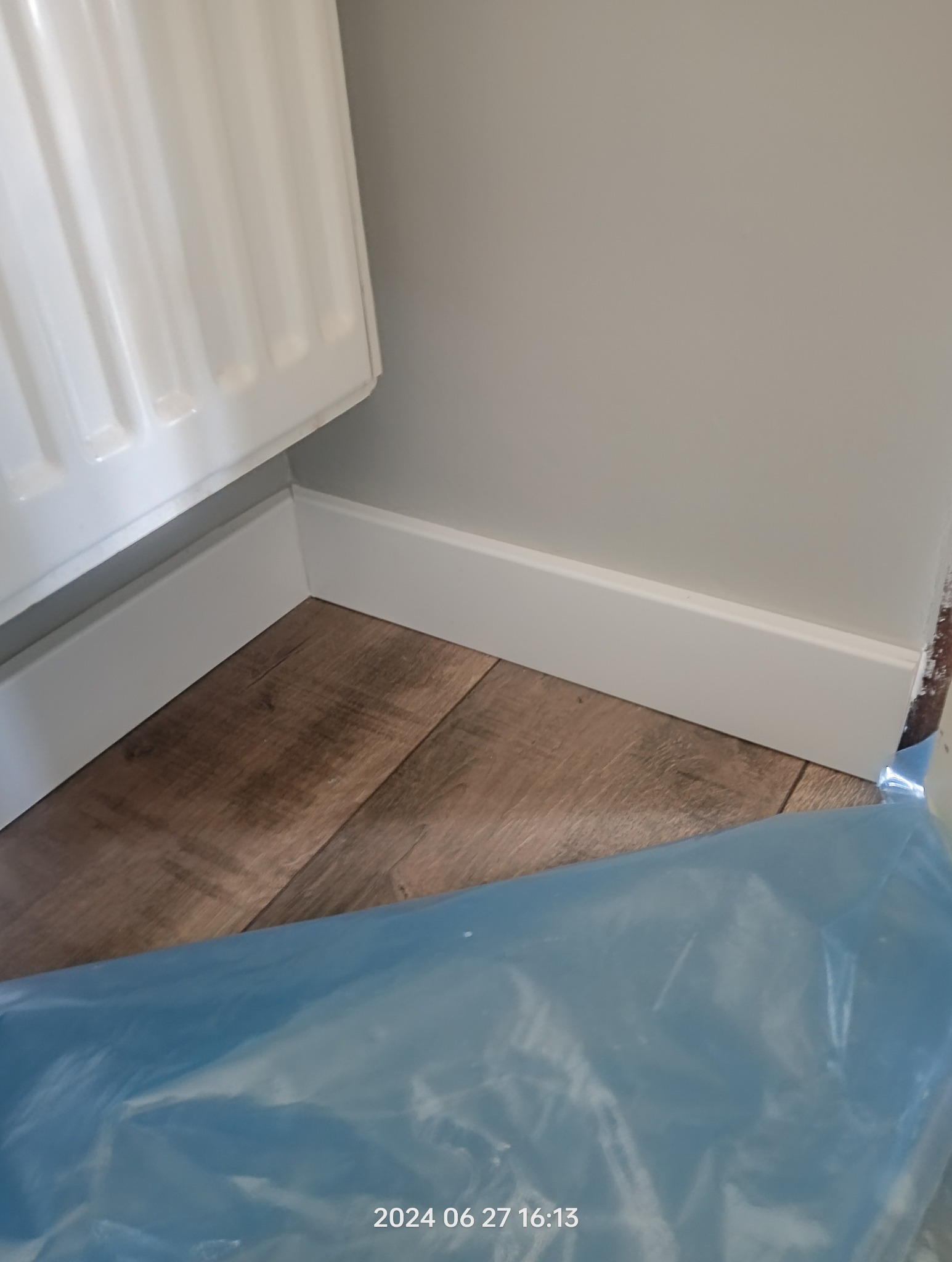 Fragment wnętrza z ułożonymi panelami podłogowymi i pomalowanymi listwami przypodłogowymi, częściowo zakryty folią ochronną.