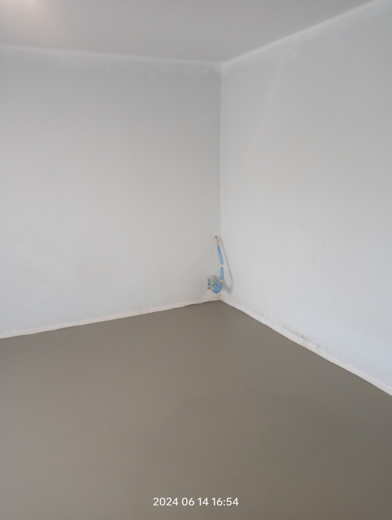 Świeżo wylana, gładka, szara wylewka samopoziomująca w rogu pomieszczenia z widocznymi rurami instalacji wodnej wystającymi ze ściany.