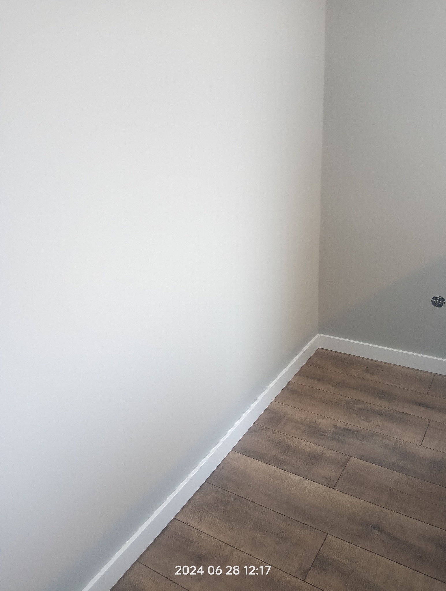 Wykończony narożnik pokoju z ułożonymi panelami podłogowymi w odcieniach brązu i pomalowanymi na biało ścianami z listwami przypodłogowymi.