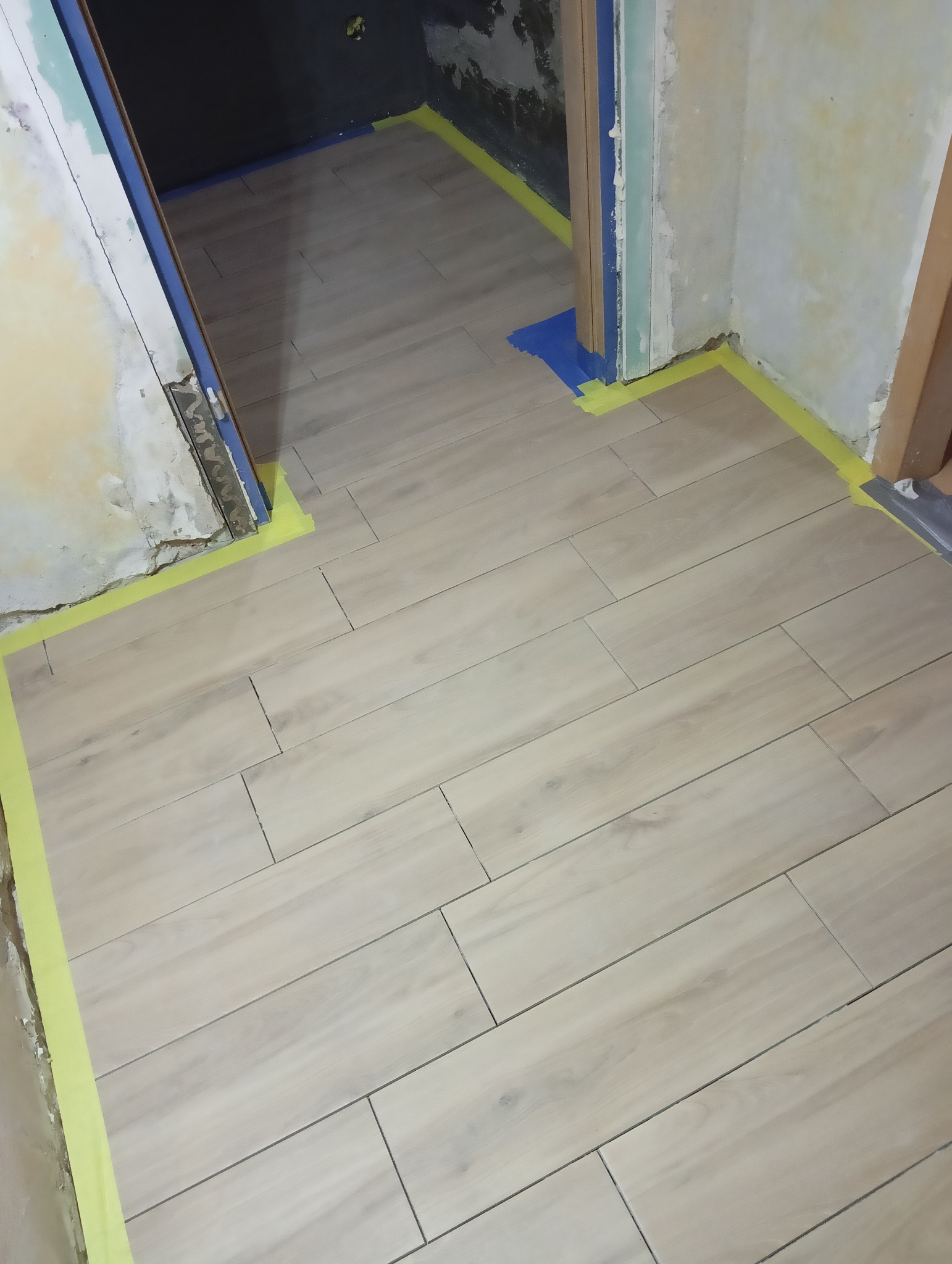 Ułożone panele podłogowe w odcieniach beżu, zabezpieczone taśmą malarską przy ścianach w trakcie remontu. Widoczne fragmenty surowych ścian i futryn.