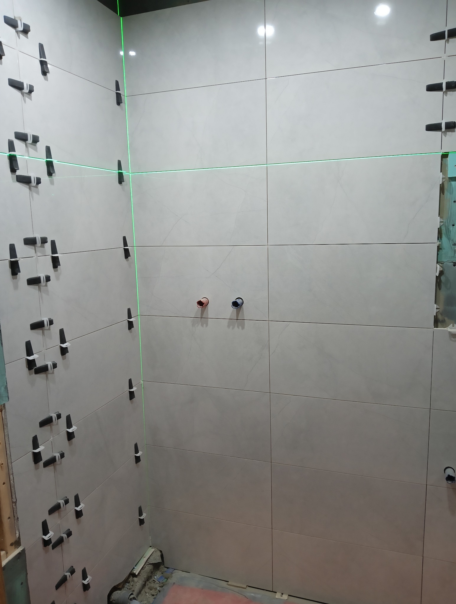 Ściany łazienki z jasnymi płytkami w trakcie montażu, widoczne klipsy poziomujące i laserowy wskaźnik, przygotowane otwory na armaturę, fragment podłogi w remoncie.