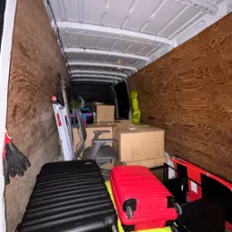 Wnętrze furgonetki wypełnione kartonami, walizkami w kolorach czarnym, czerwonym i limonkowym, krzesłem biurowym i rozłożoną drabiną, zabezpieczone drewnianą płytą.