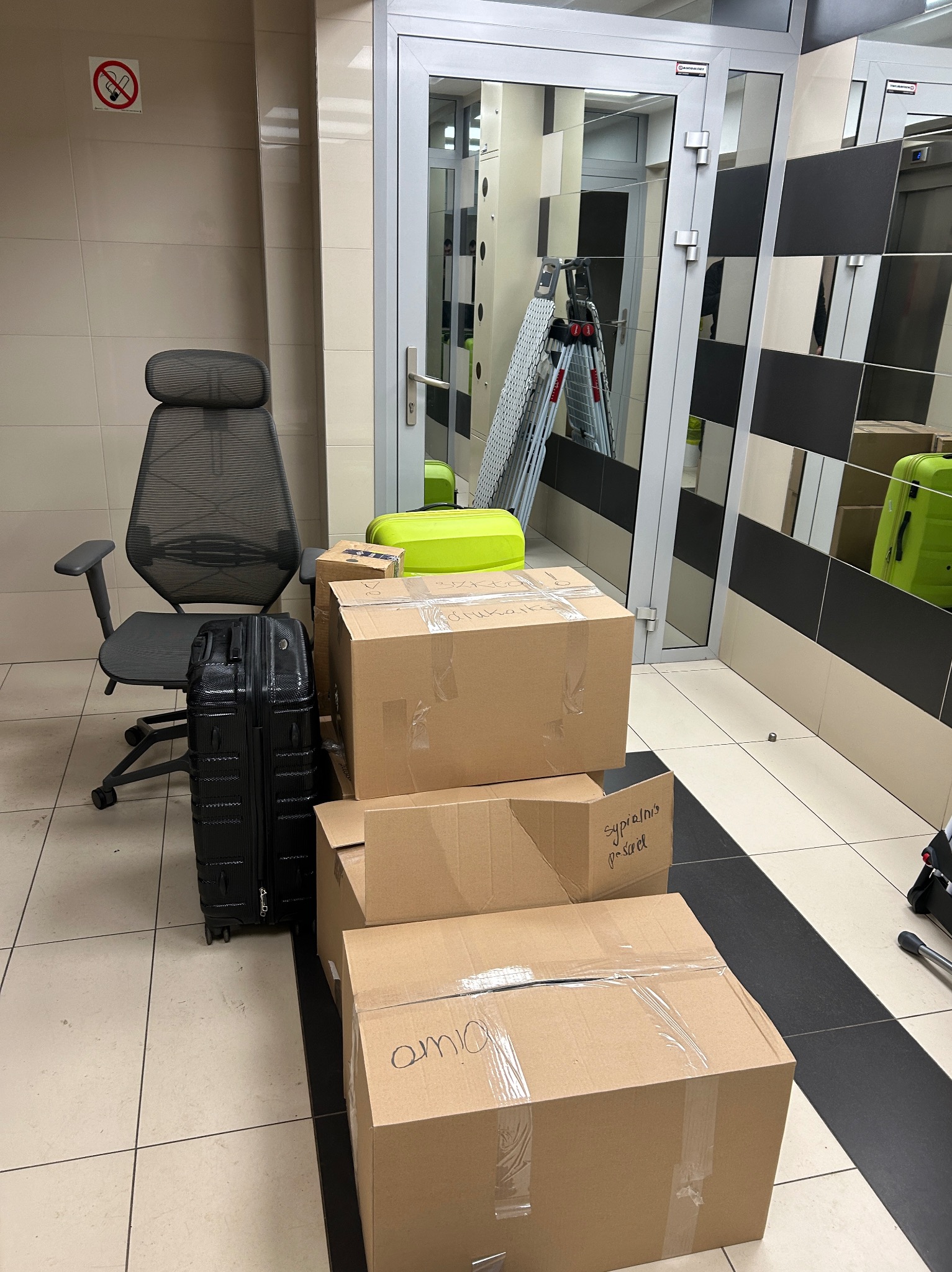 Spakowane kartony, walizka i krzesło biurowe w korytarzu przed windą, gotowe do transportu.