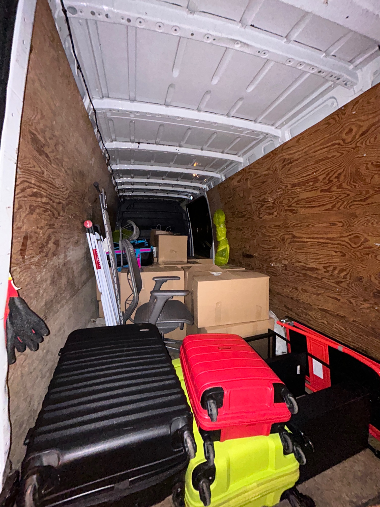 Wnętrze furgonetki wypełnione kartonami, walizkami w kolorach czarnym, czerwonym i limonkowym, krzesłem biurowym i rozłożoną drabiną, zabezpieczone drewnianą płytą.