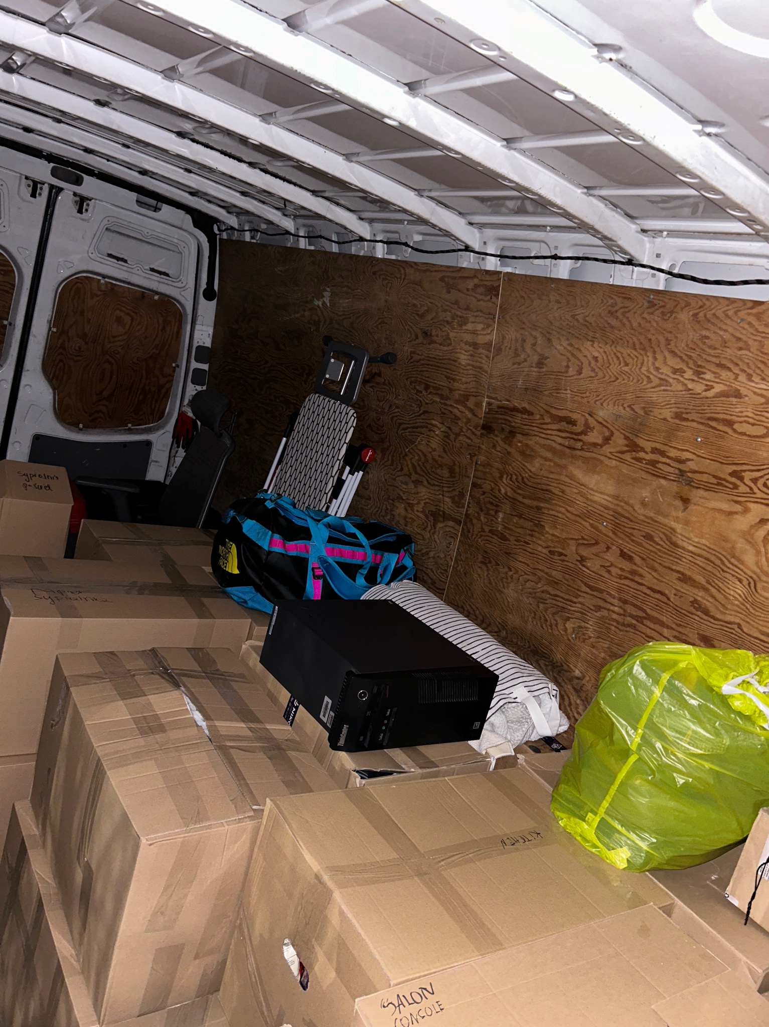 Wnętrze furgonetki wypełnione kartonami, torbą podróżną North Face, rowerem stacjonarnym i workiem na śmieci, przygotowane do transportu.
