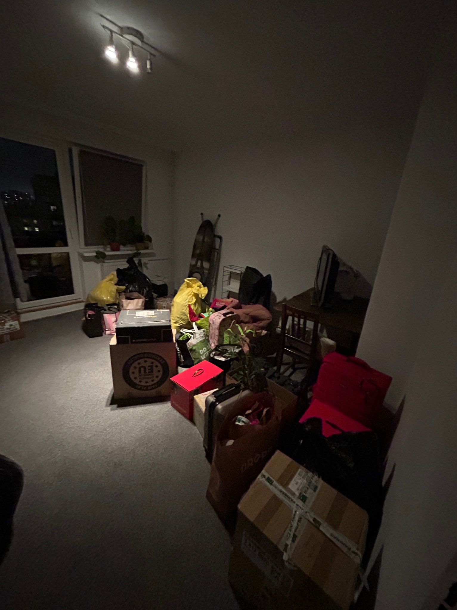 Pokój przed przeprowadzką: kartony, torby i worki z rzeczami przygotowane do transportu, widoczne meble i okno z widokiem na miasto w tle.