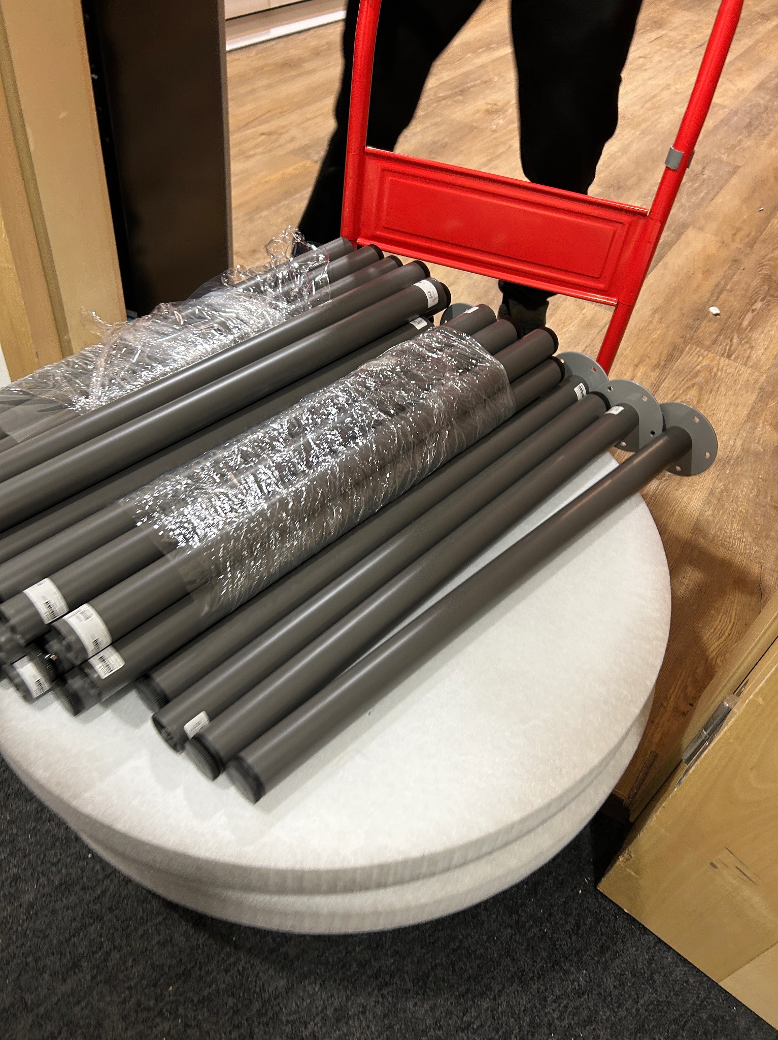Rury z metalowymi podstawami, owinięte folią, ułożone na okrągłym, białym siedzisku wózka transportowego z czerwonym uchwytem.
