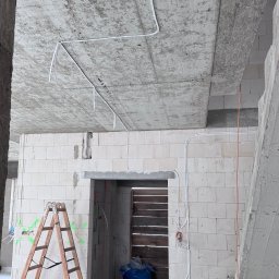 Mateusz Kalicki - Wnętrze w trakcie prac instalacyjnych: białe przewody elektryczne prowadzone po surowym, betonowym suficie i ścianach z białych bloczków. Widoczna drabina i prowizoryczne zabezpieczenie otworu.