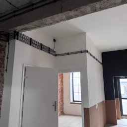 Mateusz Kalicki - Czarna instalacja elektryczna, równolegle ułożone kable przymocowane do ściany w surowym wnętrzu. Widoczne kable prowadzone wzdłuż sufitu i ścian.