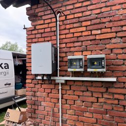 Mateusz Kalicki - Instalacja elektryczna na ceglanej ścianie budynku: falownik Sungrow, dwie skrzynki Iluve z ostrzeżeniami, rury PCV. Widoczny samochód firmy.