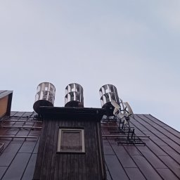 illumie - Instalacja odgromowa na dachu z blachy, widok z dołu. Trzy metalowe kominy z turbinami wiatrowymi, drabina oparta o ścianę budynku.