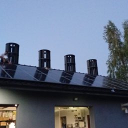 Mateusz Kalicki - Instalacja paneli słonecznych na dachu budynku z zamontowanymi odpowietrznikami. Pracownik siedzi na panelach podczas montażu. Widoczne drzewa w tle.