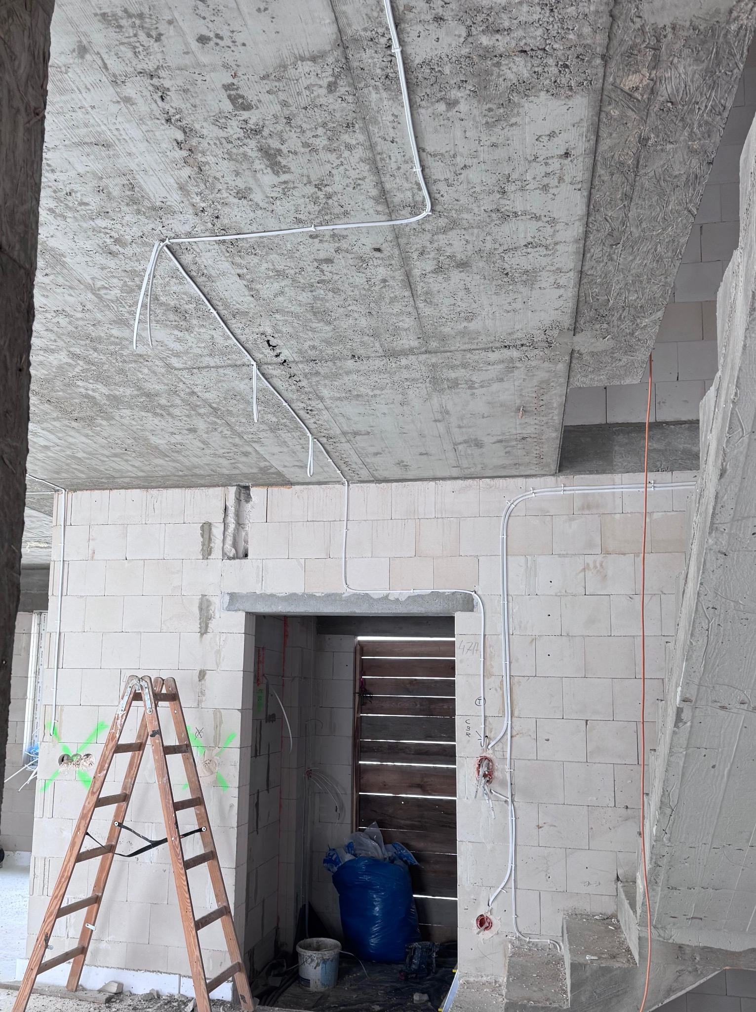 Wnętrze w trakcie prac instalacyjnych: białe przewody elektryczne prowadzone po surowym, betonowym suficie i ścianach z białych bloczków. Widoczna drabina i prowizoryczne zabezpieczenie otworu.
