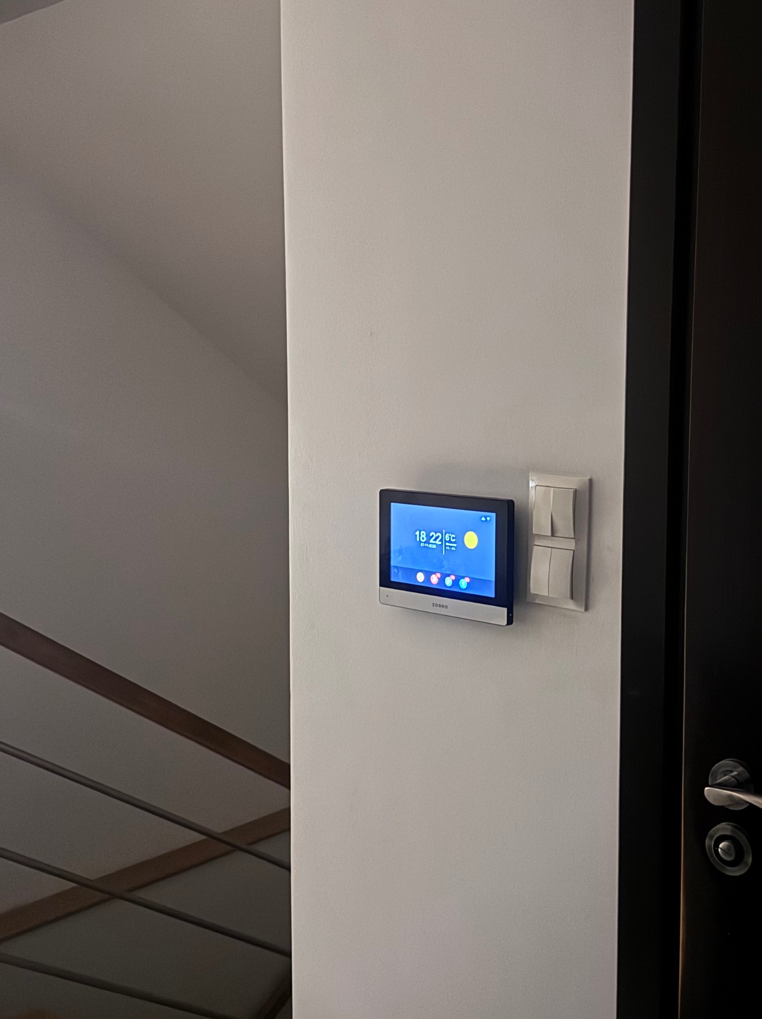 Nowoczesny panel sterowania domem inteligentnym, zamontowany na ścianie obok włącznika światła i fragmentu drzwi. Widoczny ekran z informacjami o pogodzie i godzinie.