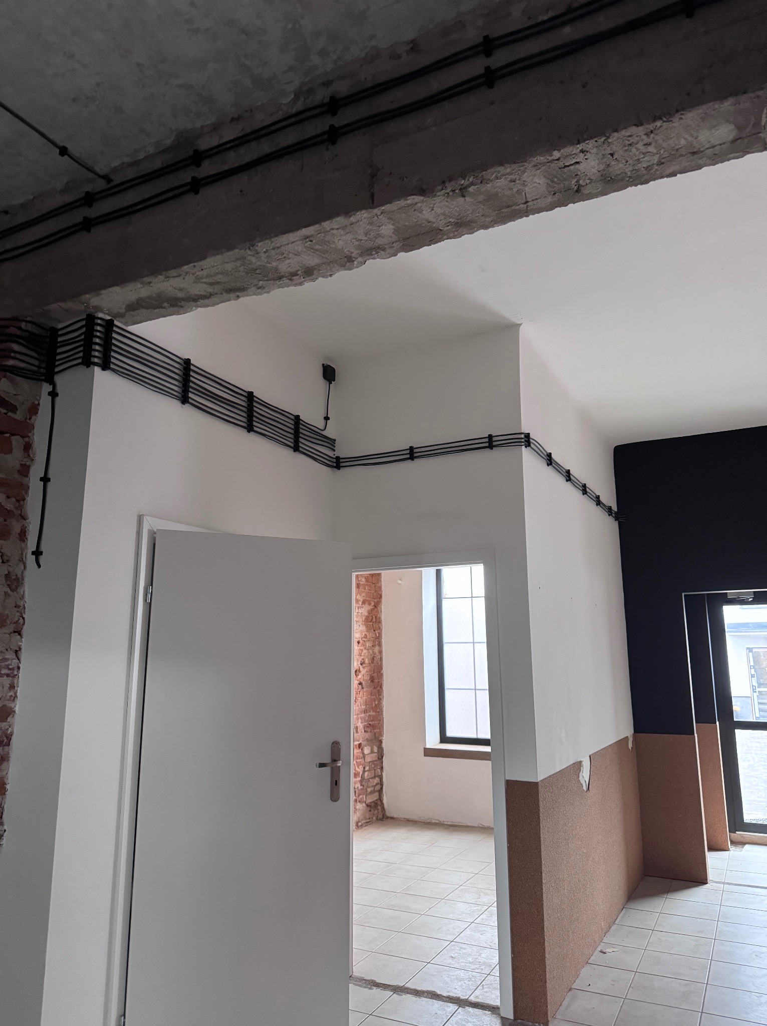 Czarna instalacja elektryczna, równolegle ułożone kable przymocowane do ściany w surowym wnętrzu. Widoczne kable prowadzone wzdłuż sufitu i ścian.