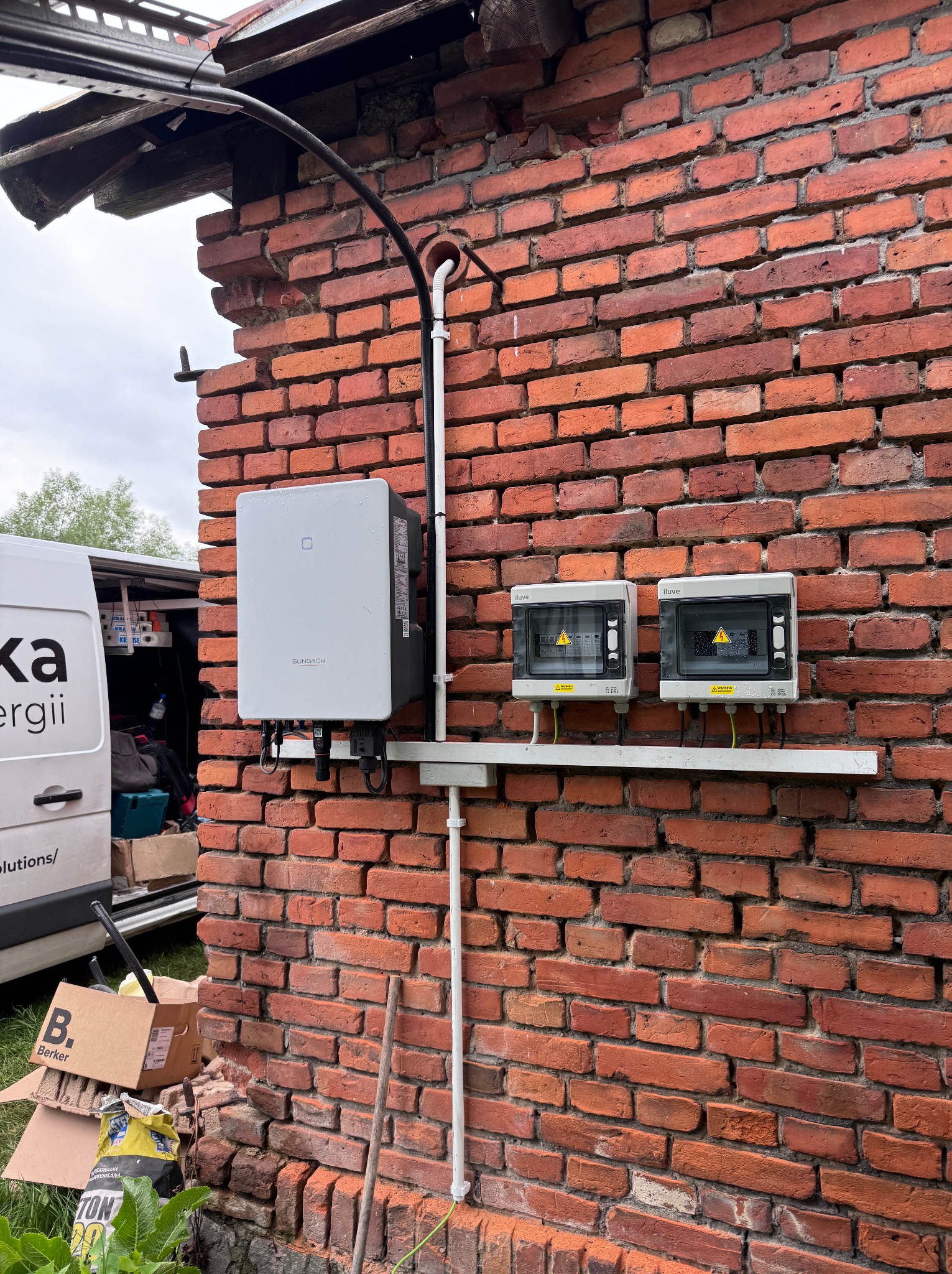 Instalacja elektryczna na ceglanej ścianie budynku: falownik Sungrow, dwie skrzynki Iluve z ostrzeżeniami, rury PCV. Widoczny samochód firmy.