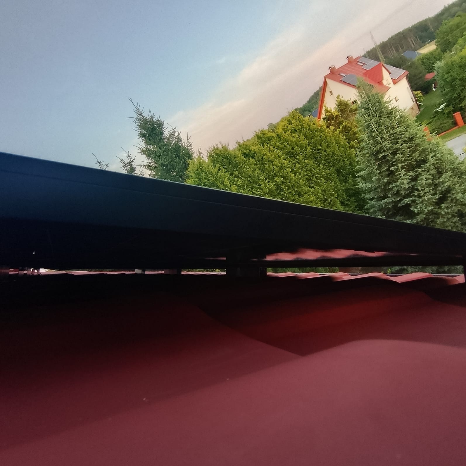 Czerwony dach z panelami słonecznymi, widok z dołu na tle zieleni i domu z czerwoną dachówką. Nietypowy kadr.