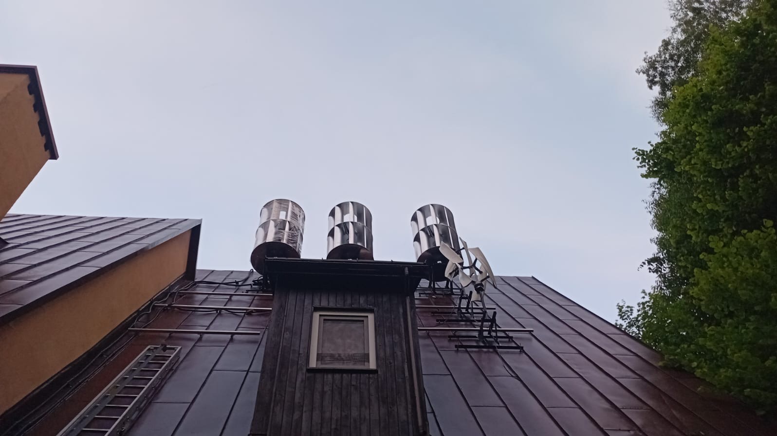 Instalacja odgromowa na dachu z blachy, widok z dołu. Trzy metalowe kominy z turbinami wiatrowymi, drabina oparta o ścianę budynku.