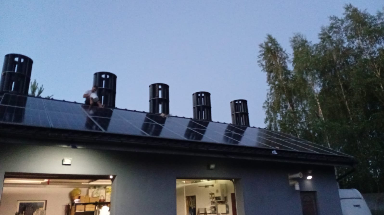 Instalacja paneli słonecznych na dachu budynku z zamontowanymi odpowietrznikami. Pracownik siedzi na panelach podczas montażu. Widoczne drzewa w tle.