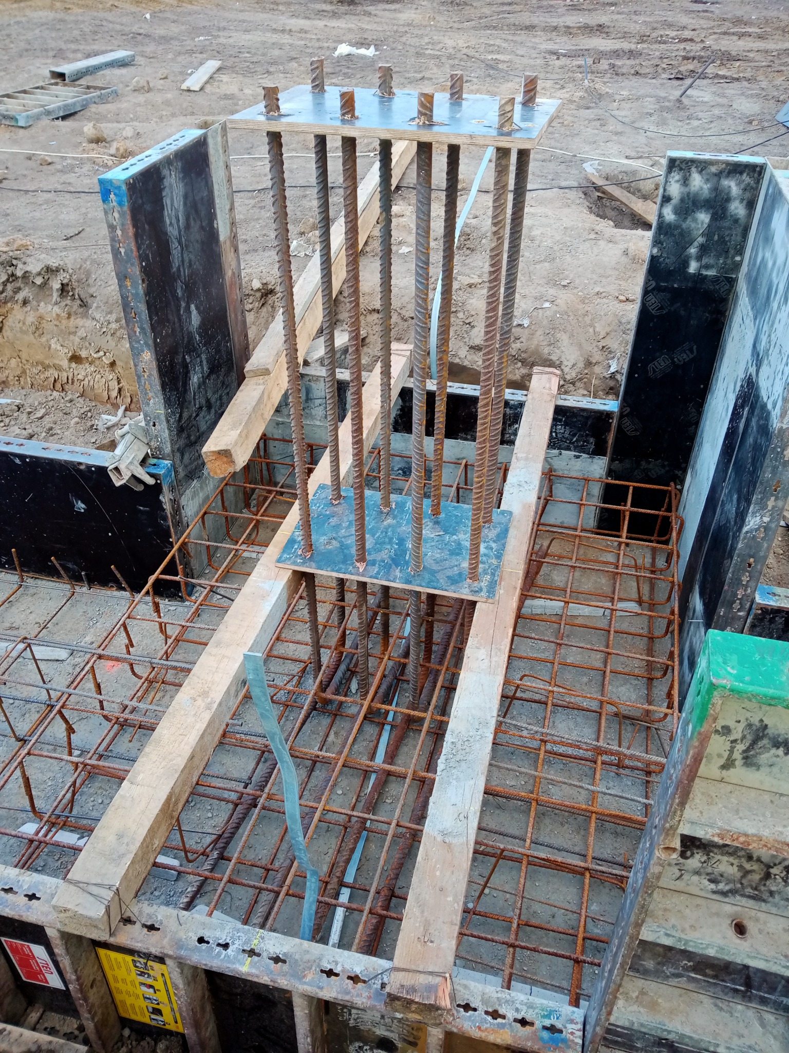 Zbrojenie fundamentu z wystającymi prętami pionowymi, szalunek drewniany i stalowy, widoczne z góry na placu budowy.
