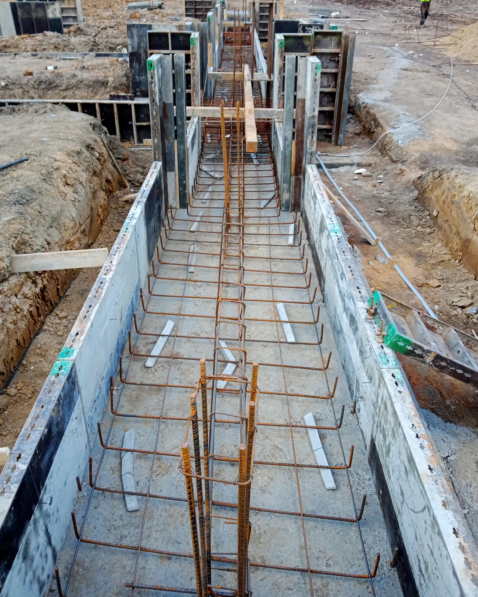 Szalunek i zbrojenie pod wylewkę fundamentów, widoczne stalowe pręty zbrojeniowe i betonowe elementy szalunku, ziemia wokół wykopu.