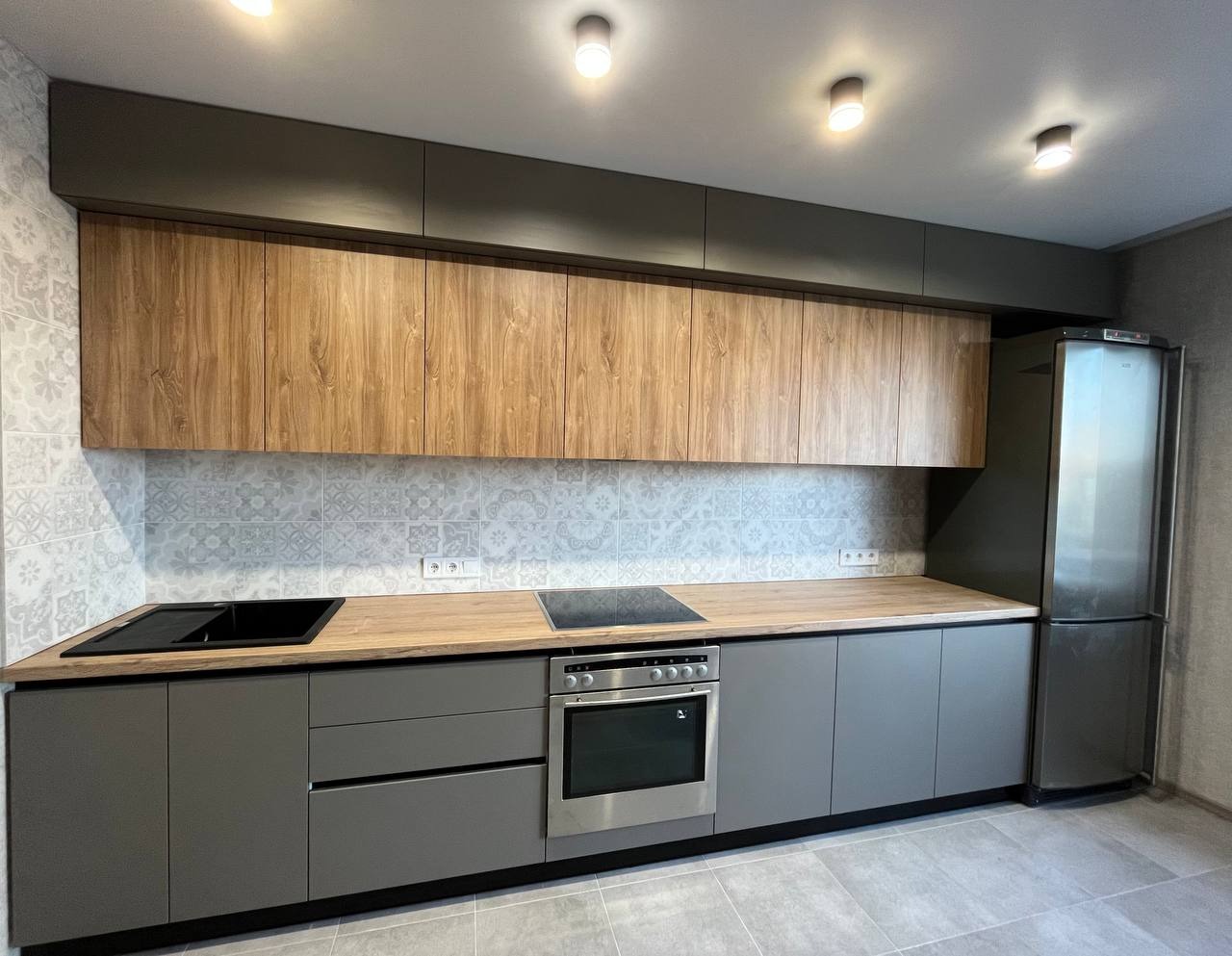 Nowoczesna kuchnia z szarymi dolnymi szafkami, drewnianymi górnymi szafkami, blatem w kolorze drewna i płytkami w stylu patchwork na ścianie.