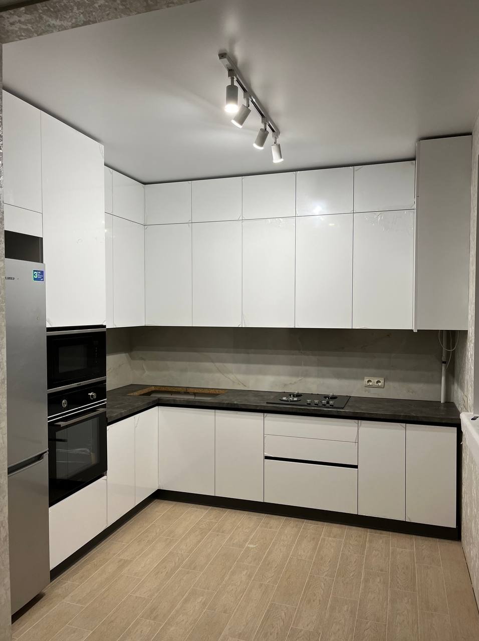 Biała, nowoczesna kuchnia z czarnym blatem, widoczne oświetlenie punktowe na suficie i sprzęt AGD w zabudowie.
