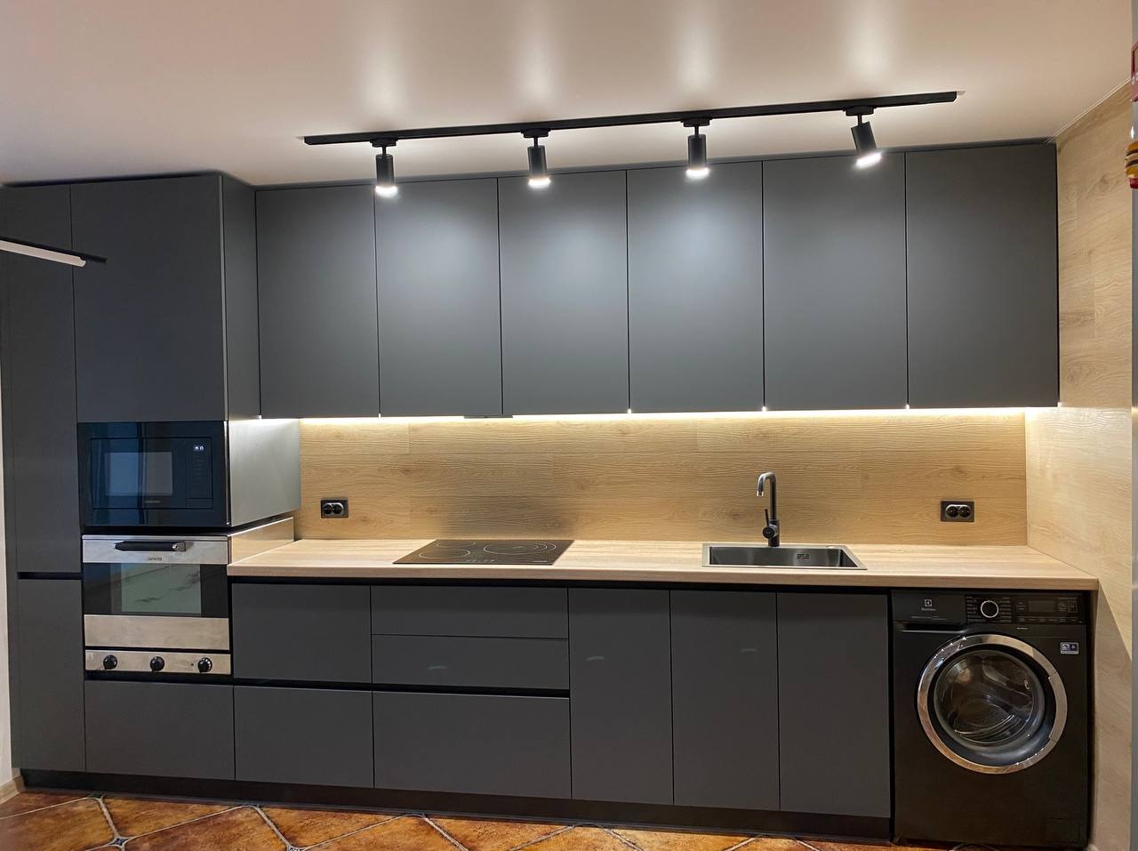 Szara zabudowa kuchenna z oświetleniem punktowym i drewnianym panelem ściennym, integrująca piekarnik, mikrofalówkę, płytę indukcyjną, zlew i pralkę.