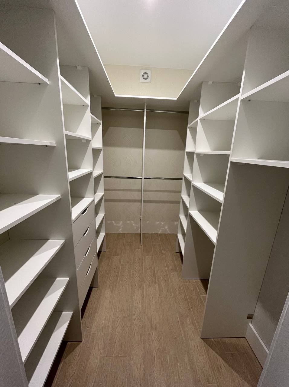 Wnętrze garderoby z białymi regałami i szufladami, oświetlenie sufitowe, drewnopodobna podłoga.