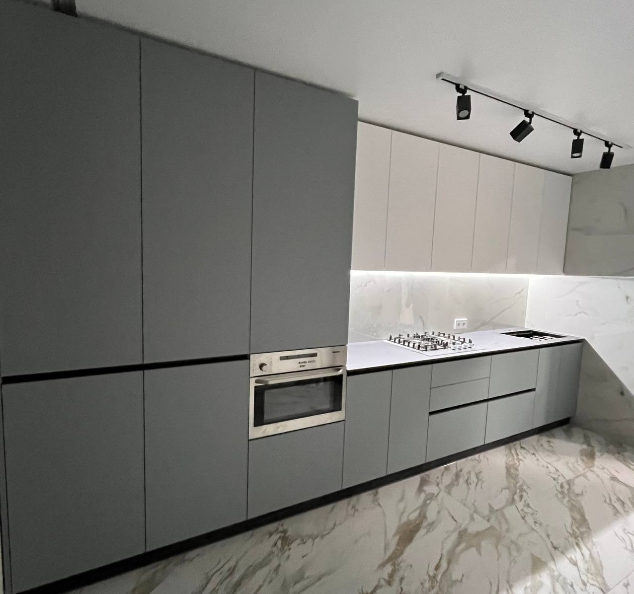 Nowoczesna kuchnia z szarymi matowymi frontami, wbudowanym piekarnikiem i marmurowym blatem oraz podświetleniem LED pod szafkami wiszącymi.
