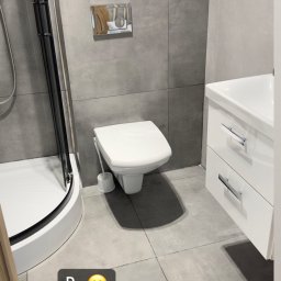 My clean team - Wyremontowana łazienka z szarą glazurą, wiszącą toaletą, kabiną prysznicową z czarnymi ramami i białym zlewem z szafkami, widoczny bojler nad mozaiką.